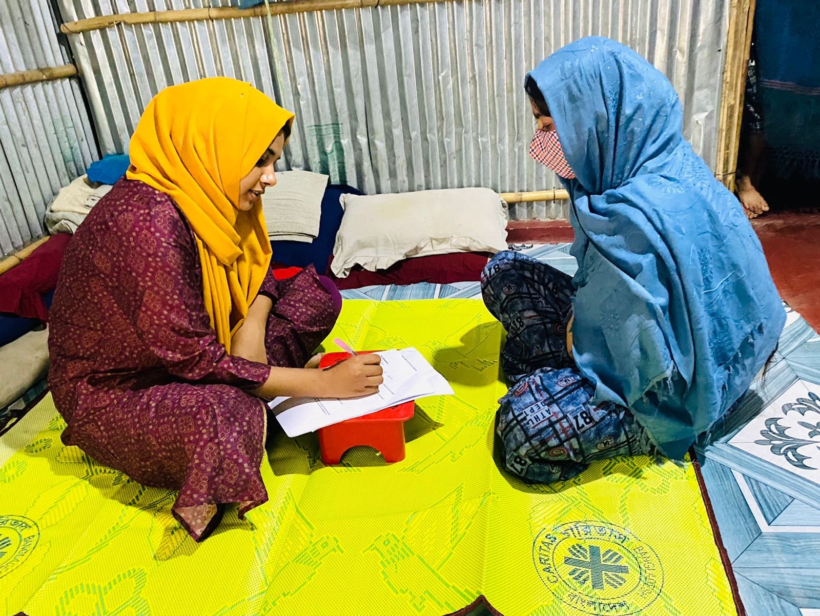 Two women sat on the floor in a camp for Rohingya people - talking about health care barriers